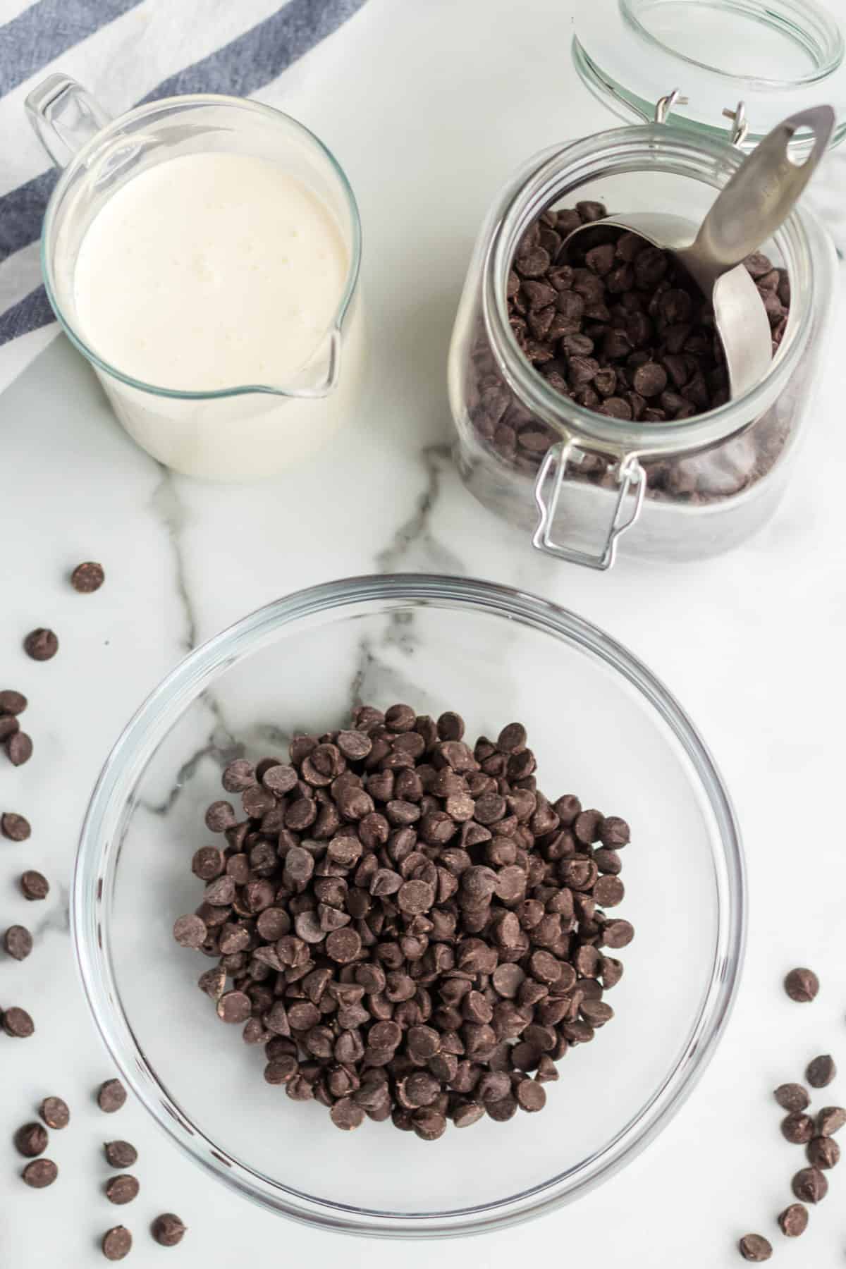 A glass bowl filled with chocolate chips, a few scattered on a marble surface, sits beside a jar of chocolate chips with a metal scoop and a glass measuring cup of cream—perfect ingredients for your next chocolate mousse recipe.
