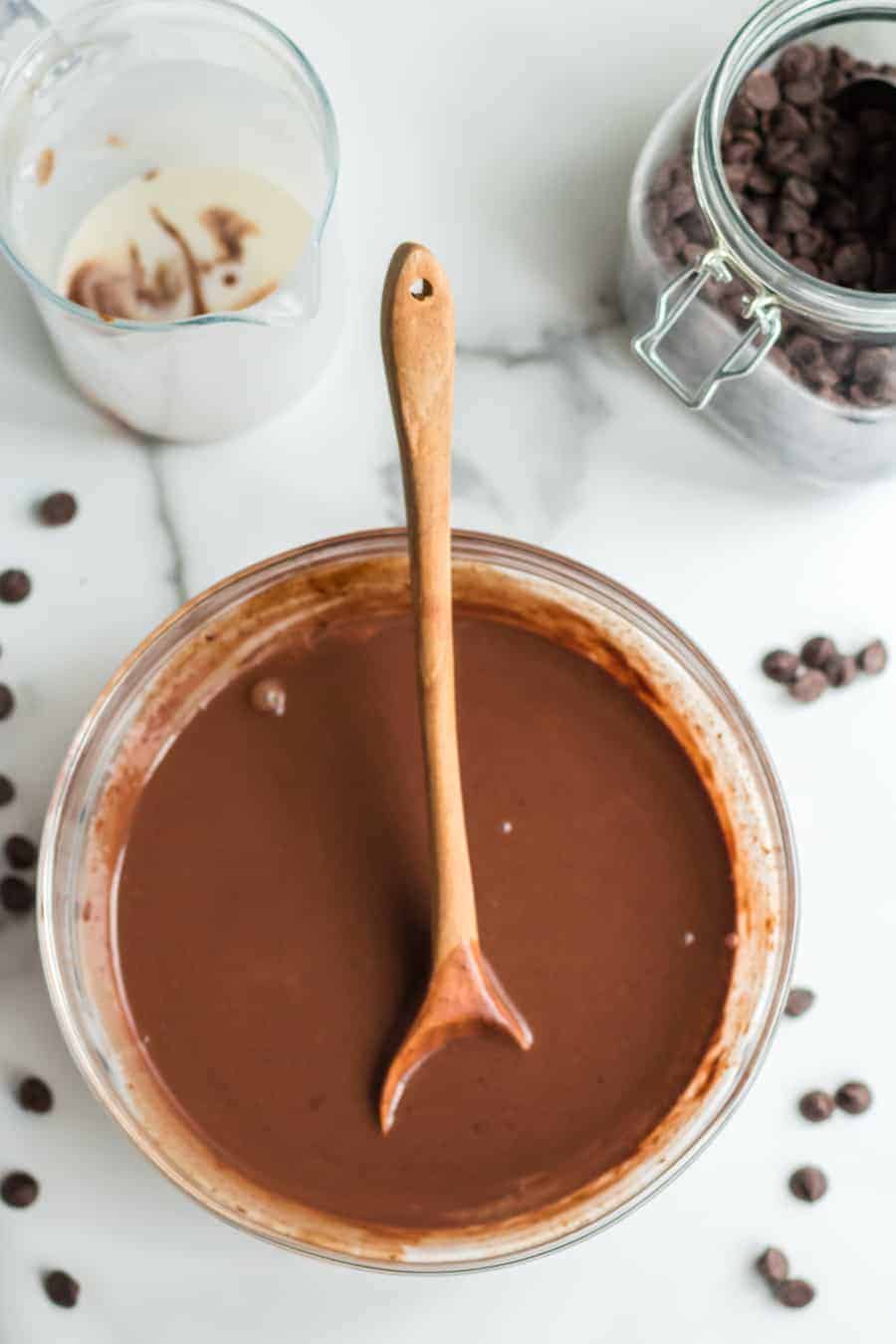 A glass bowl filled with chocolate batter and a wooden spoon, surrounded by scattered chocolate chips—perfect for starting your favorite chocolate mousse recipe. Nearby are a glass jar of chocolate chips and a pitcher with milk and chocolate residue.