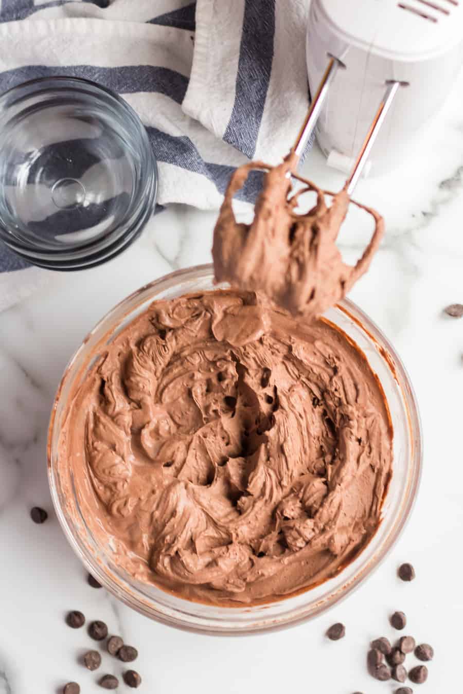 A glass bowl filled with whipped chocolate mousse, made from a rich chocolate mousse recipe, sits on a marble surface. A hand mixer with mousse-coated beaters rests above the bowl, surrounded by chocolate chips and a glass cup.