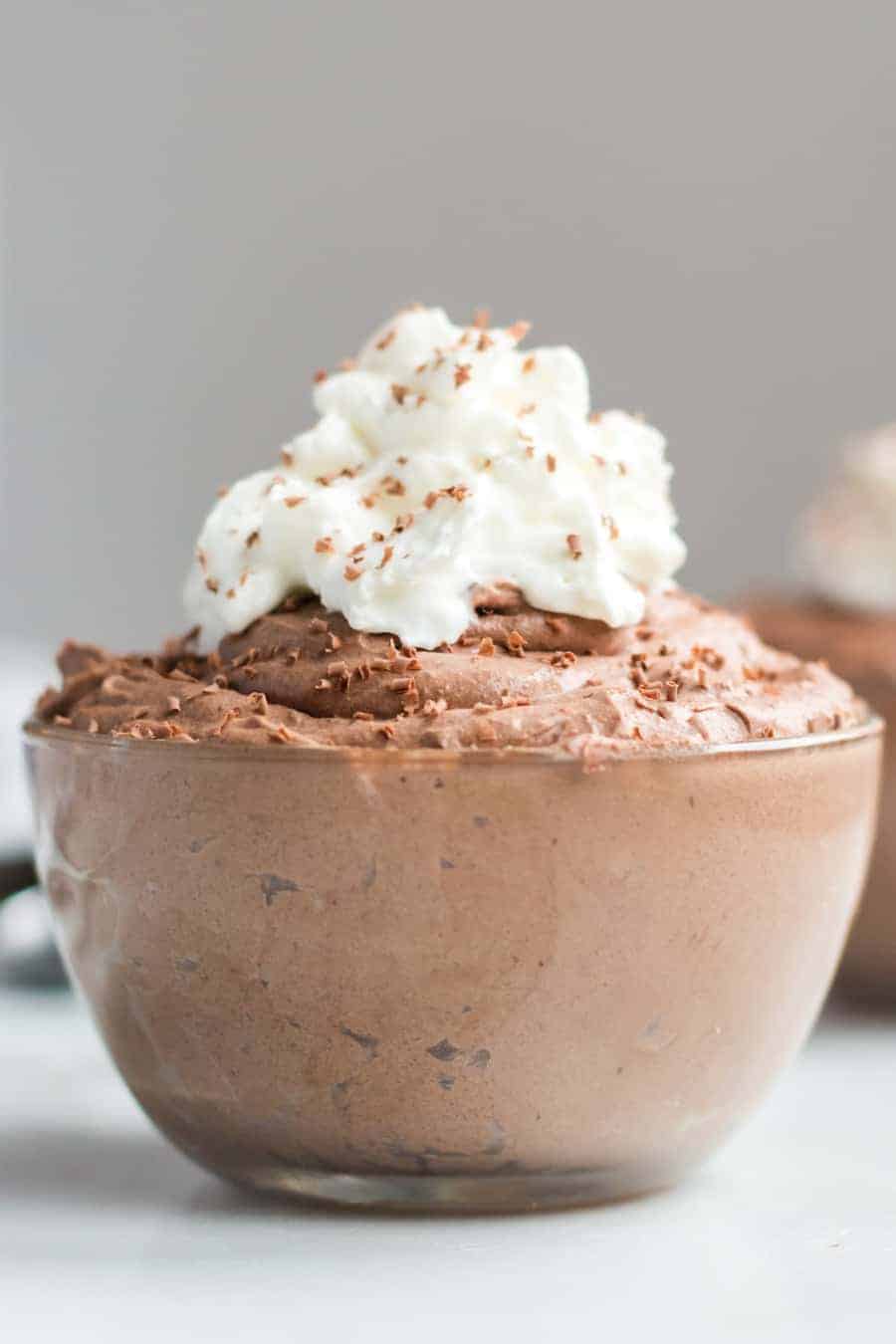 A glass bowl filled with creamy chocolate mousse, made from a rich chocolate mousse recipe, topped with a swirl of whipped cream and chocolate shavings, set against a light, blurred background.