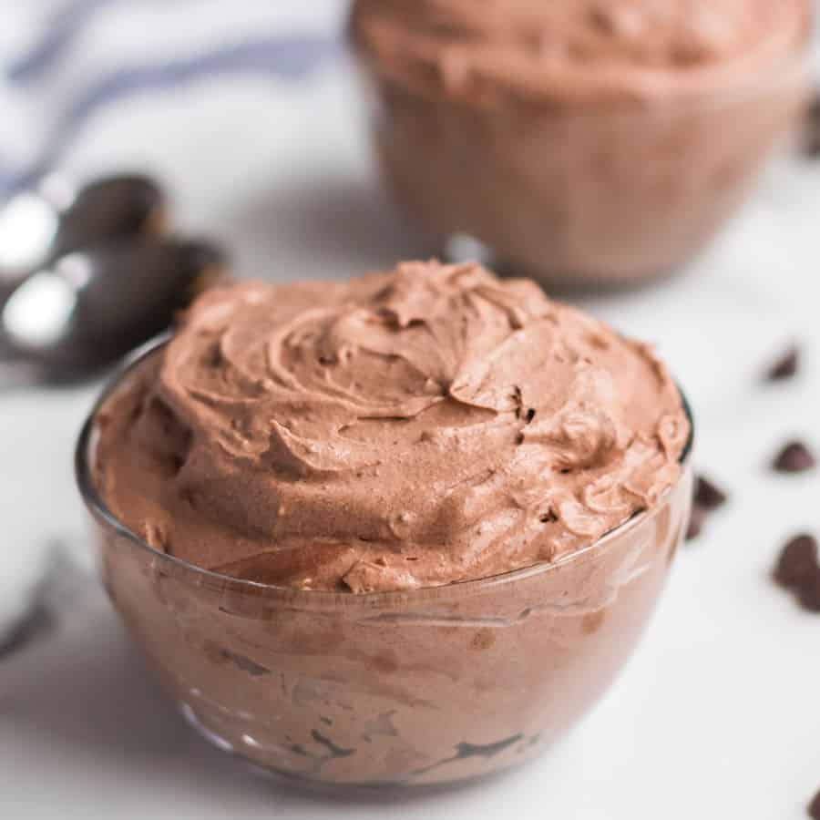 A glass bowl filled with creamy chocolate mousse sits on a white surface, hinting at an irresistible chocolate mousse recipe, with another bowl and two spoons in the background, plus scattered chocolate pieces.