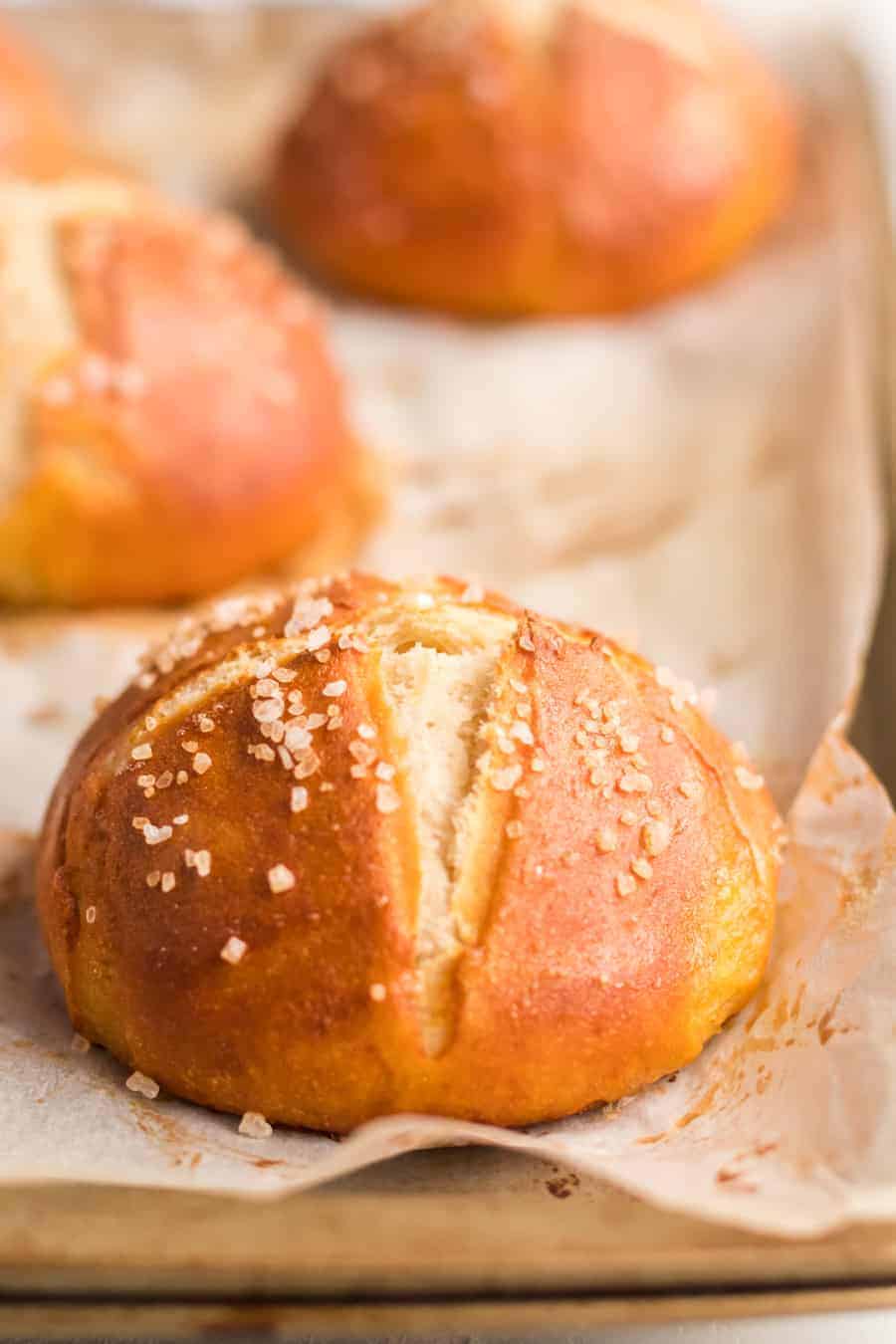 A close-up of a golden-brown pretzel roll topped with coarse salt sits on parchment paper. More rolls from this delicious pretzel rolls recipe are visible in the background, slightly out of focus.