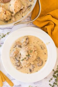 A white plate with creamy chicken and mushrooms from a classic chicken fricassee recipe, garnished with fresh thyme, sits on marble next to golden utensils, a yellow napkin, and a pot filled with more of the savory dish.