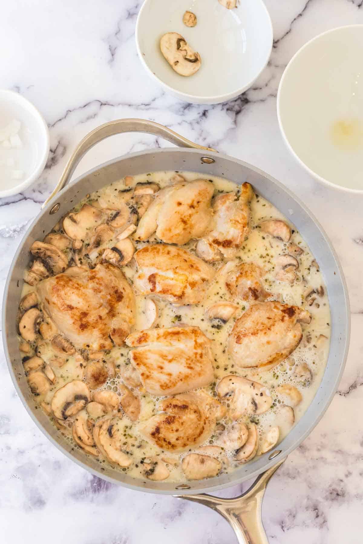 A skillet filled with cooked chicken breasts and sliced mushrooms in a creamy sauce, showcasing a delicious chicken fricassee recipe. Nearby are two small bowls, one with mushroom slices and the other mostly empty.