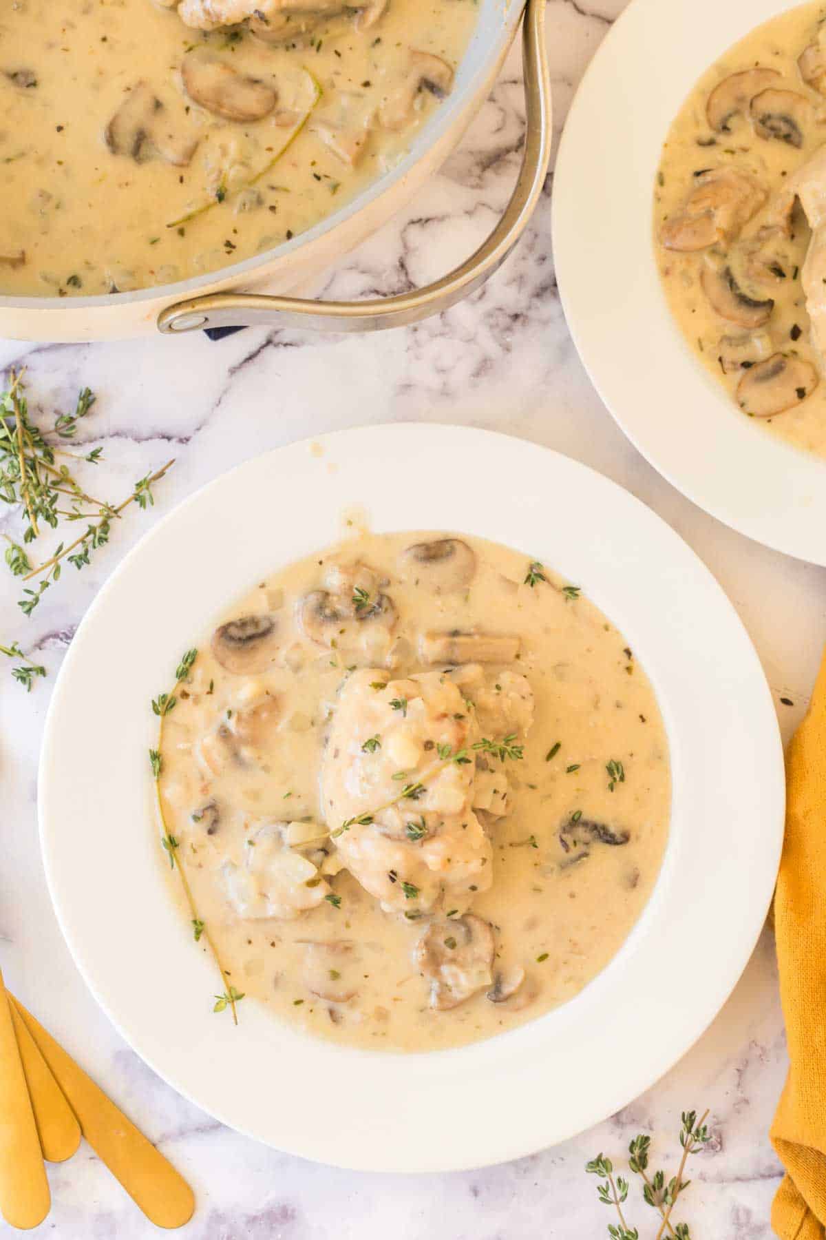 A white plate with chicken fricassee recipe in a creamy mushroom sauce, garnished with fresh thyme, sits on a marble surface near a pot of the same dish and an orange napkin with gold utensils.