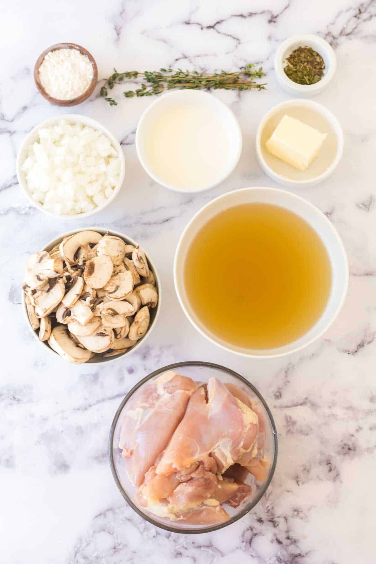 A marble countertop displays ingredients for a chicken fricassee recipe: raw chicken, sliced mushrooms, chopped onion, grated parmesan, broth, cream, butter, dried herbs, and fresh thyme arranged neatly in bowls.