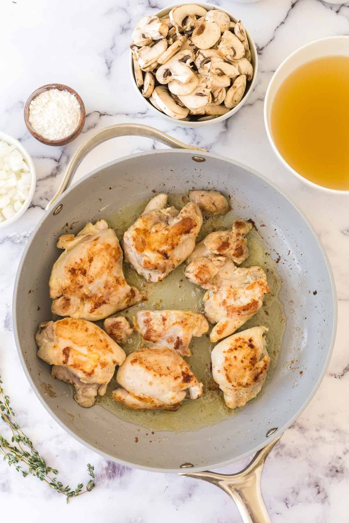A skillet with browned chicken thighs sits on a marble surface, ready for a delicious chicken fricassee recipe, surrounded by bowls of sliced mushrooms, chopped onions, grated cheese, broth, and fresh thyme sprigs.