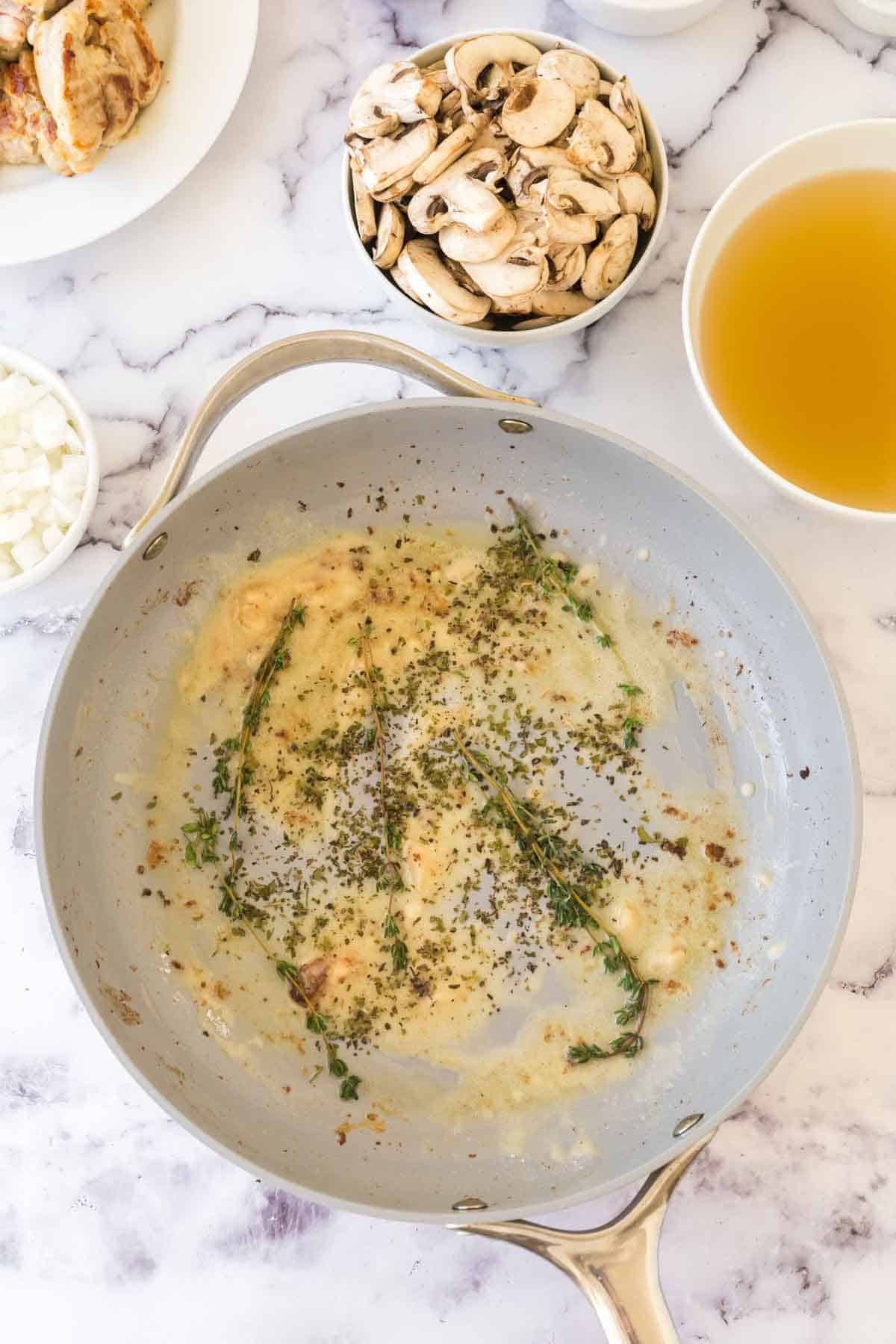 A skillet with sautéed onions, herbs, and garlic sits on a marble countertop, surrounded by bowls of sliced mushrooms, diced onions, chicken broth, and a plate of cooked chicken—perfect ingredients for a classic chicken fricassee recipe.