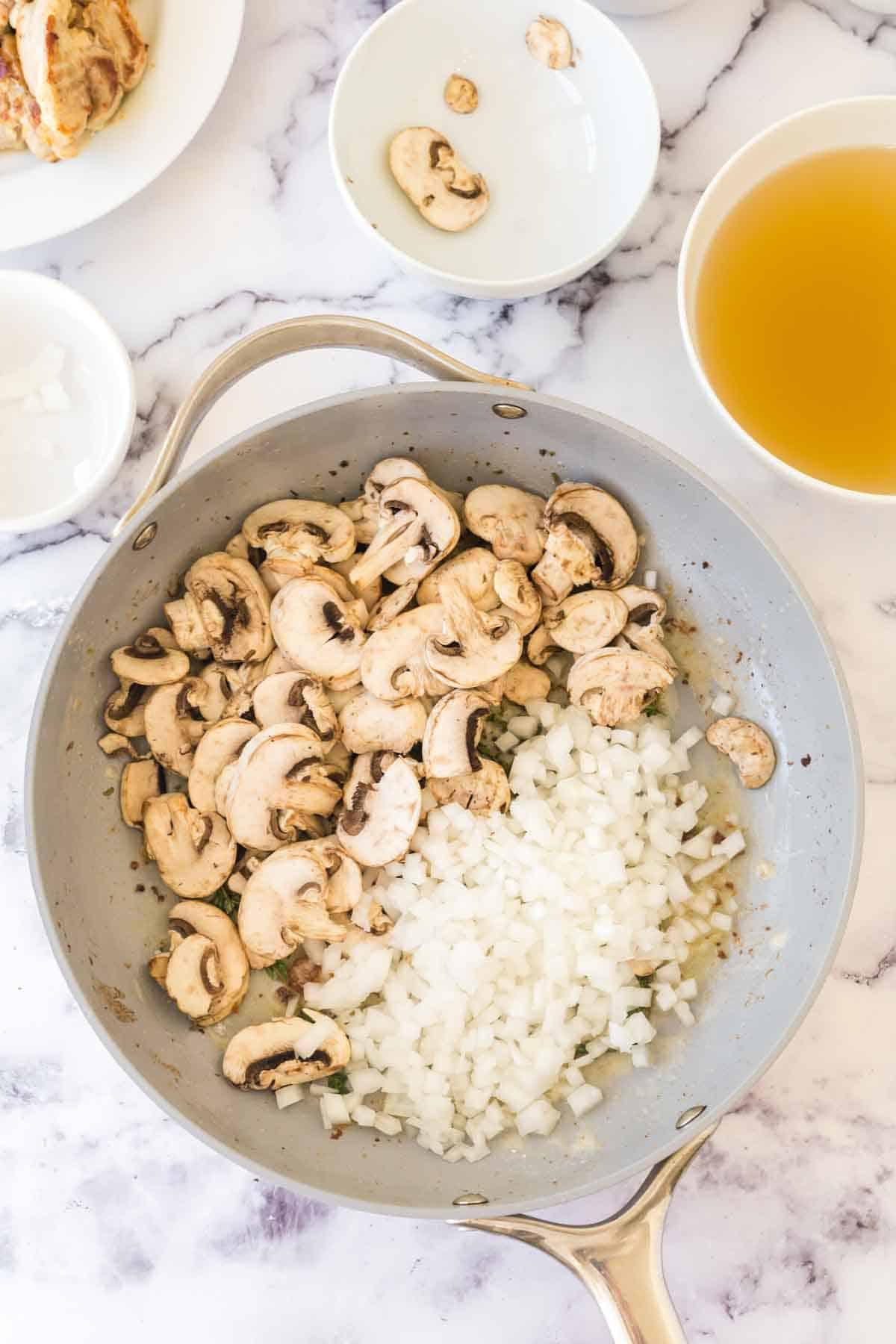 A frying pan with sliced mushrooms and chopped onions cooking, placed on a marble countertop. Surrounding the pan are small bowls containing broth, mushroom slices, and additional onion—perfect ingredients for a classic chicken fricassee recipe.