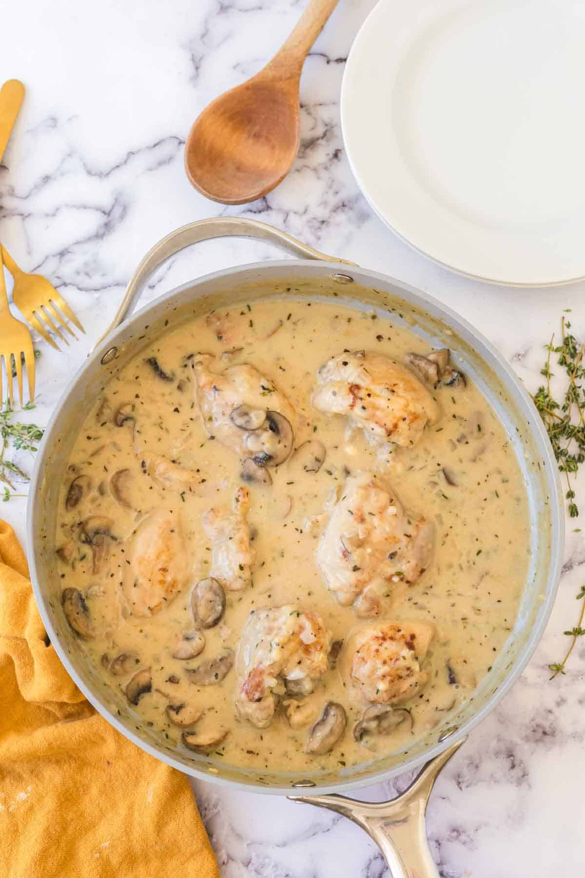 A skillet filled with chicken thighs in a creamy mushroom sauce, inspired by a classic chicken fricassee recipe, sits on a marble surface surrounded by a wooden spoon, white plate, gold fork and knife, fresh thyme, and a yellow cloth.