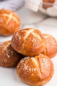 A close-up of four golden-brown pretzel rolls stacked on a white surface, topped with coarse salt, showcases the delicious result of a classic pretzel rolls recipe, with another roll and a basket blurred in the background.