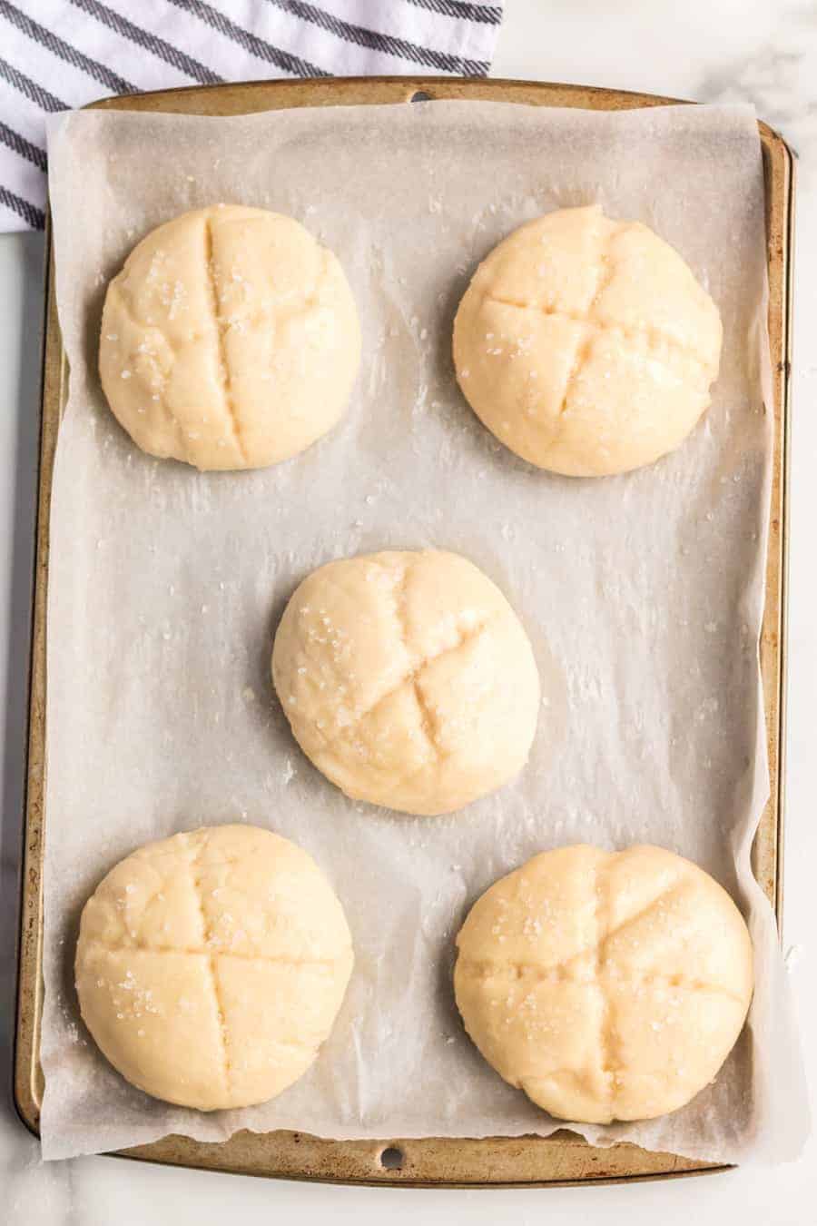 Five unbaked, round dough balls with cross-shaped cuts on top are arranged on a parchment-lined baking sheet, ready for the oven—an inviting start to any pretzel rolls recipe. A striped towel is partially visible in the corner.