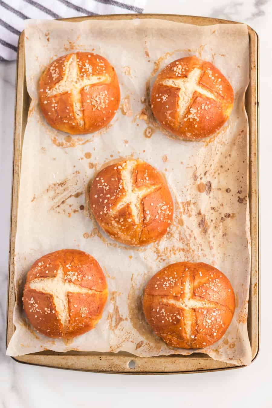 Five golden-brown pretzel rolls with salted tops and cross-shaped slashes are arranged on a parchment-lined baking tray. A striped towel is partially visible in the top left corner, hinting at a fresh batch from your favorite pretzel rolls recipe.