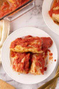 A white plate with several cabbage rolls from a classic stuffed cabbage rolls recipe, covered in tomato sauce, set on a marble surface next to utensils and a baking dish.
