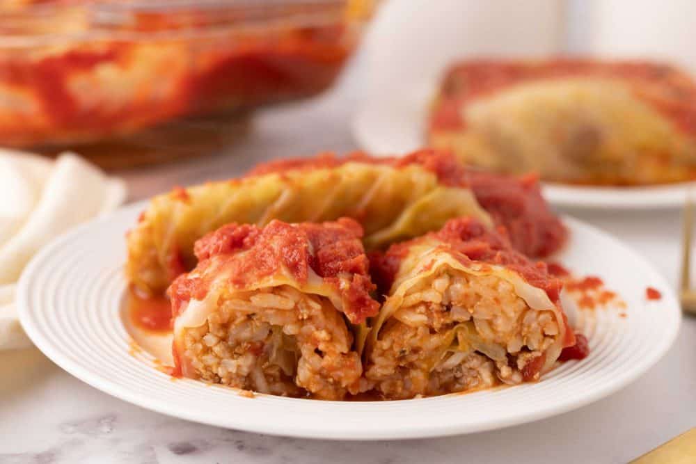 A white plate with stuffed cabbage rolls filled with rice and meat, topped with tomato sauce. One roll is cut open to reveal the filling. In the background, a serving dish displays more of this delicious stuffed cabbage rolls recipe.