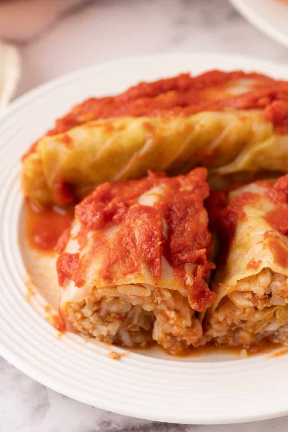 A close-up of a stuffed cabbage rolls recipe topped with tomato sauce on a white plate, with one roll cut in half to show the savory rice and meat filling inside.