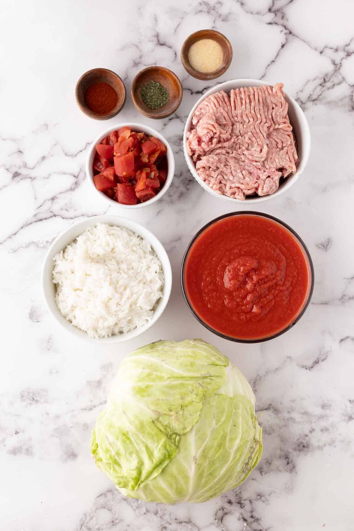 Overhead view of ingredients on a marble surface for a stuffed cabbage rolls recipe: a whole cabbage, bowls of white rice, canned tomatoes, tomato sauce, raw ground meat, and small bowls of various spices.