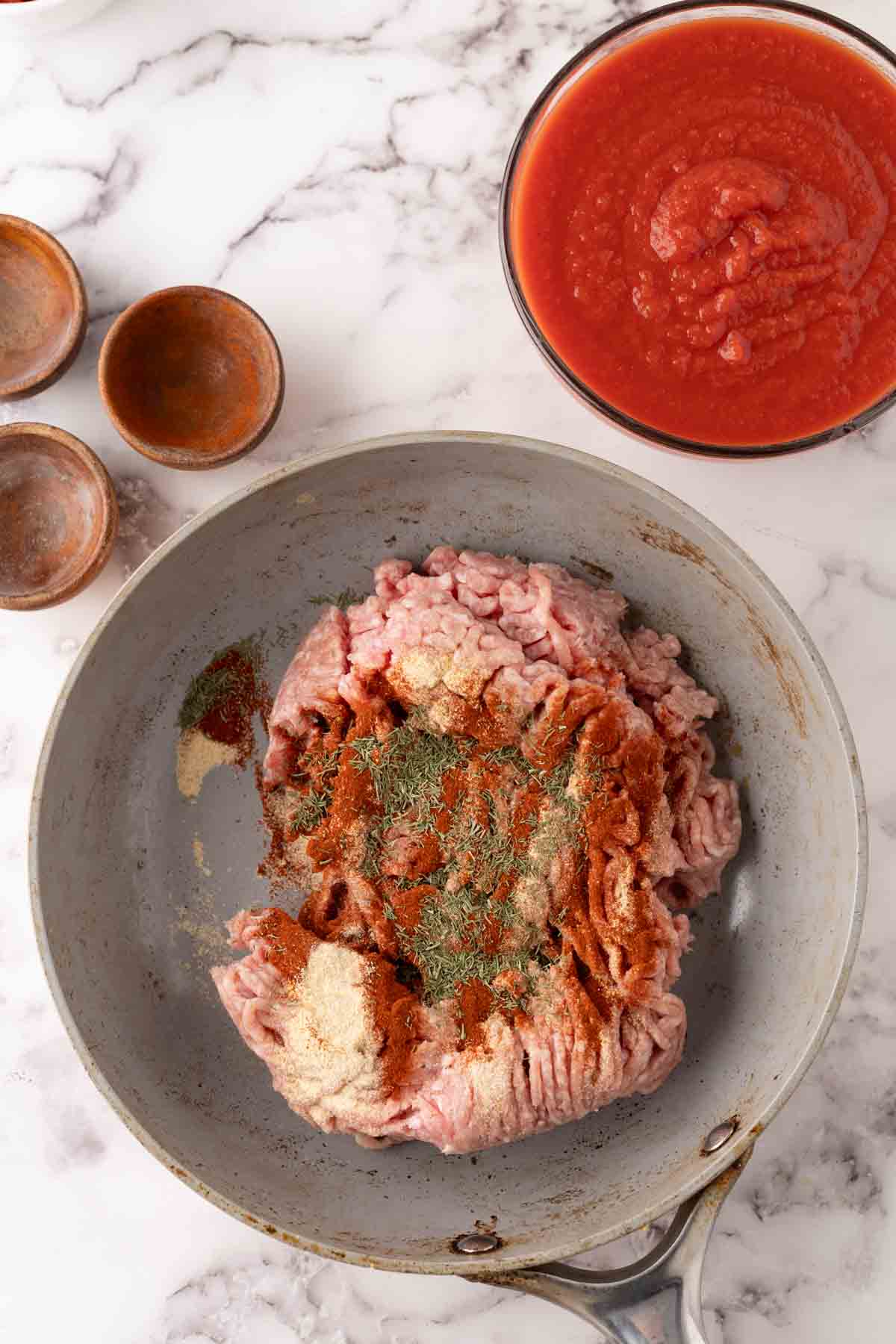 A frying pan with raw ground meat topped with various spices sits on a marble surface, ready for a stuffed cabbage rolls recipe. Nearby are a bowl of tomato sauce and three small empty wooden bowls.