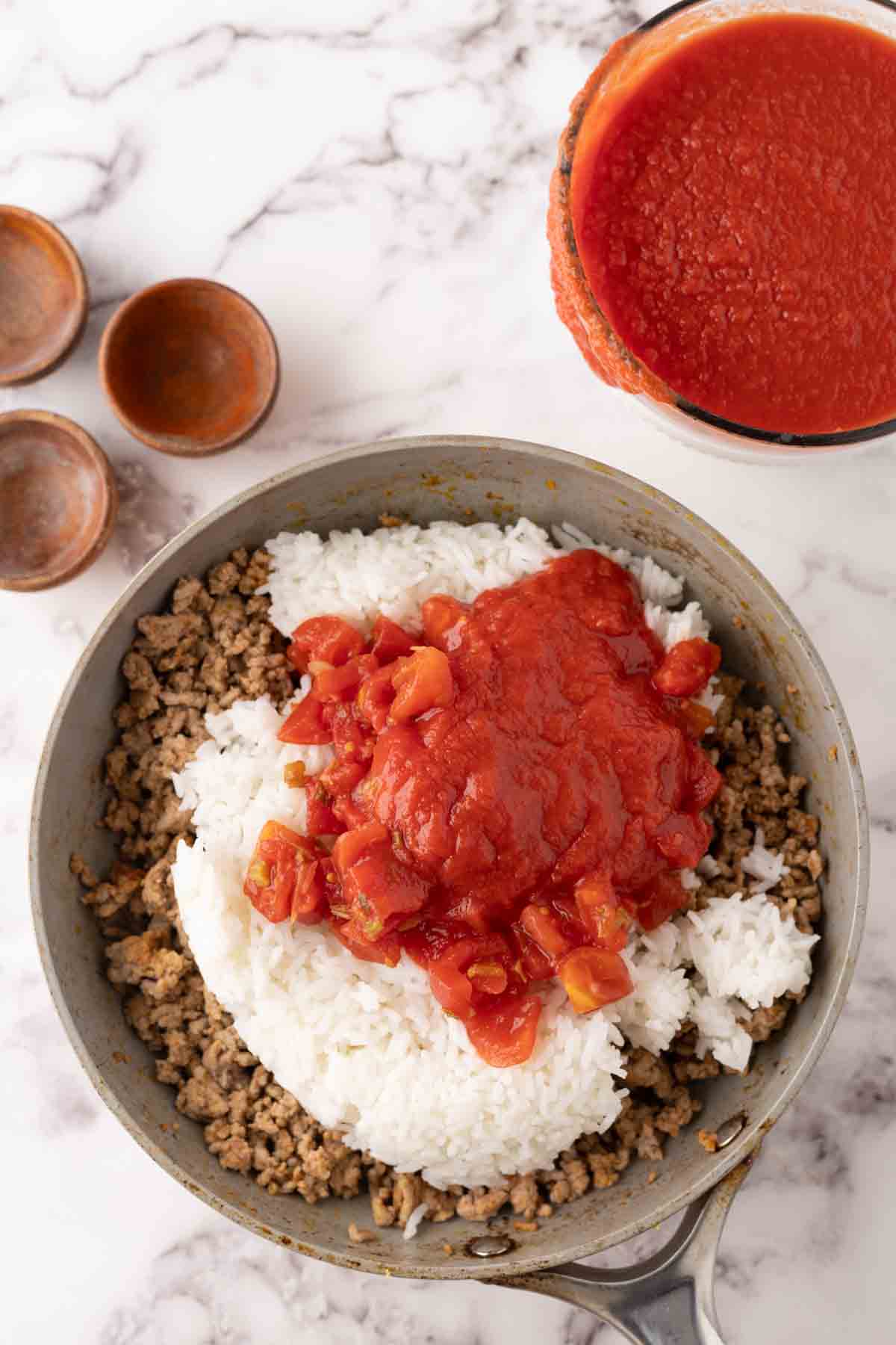 A pan with cooked ground beef, white rice, and chunky tomato sauce—ingredients often found in a classic stuffed cabbage rolls recipe—sits on a marble surface. Nearby are a bowl of extra tomato sauce and three small empty bowls.