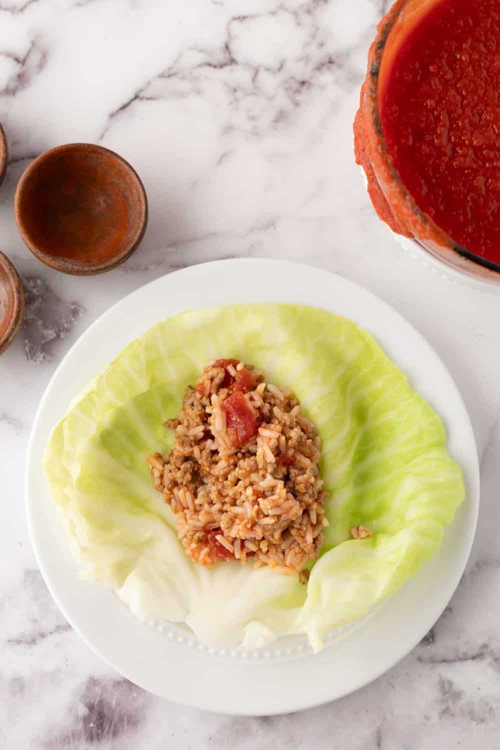 A cooked cabbage leaf on a white plate filled with a savory mixture of rice, tomatoes, and ground meat—this classic stuffed cabbage rolls recipe is served alongside a bowl of red tomato sauce and empty bowls on a marble surface.