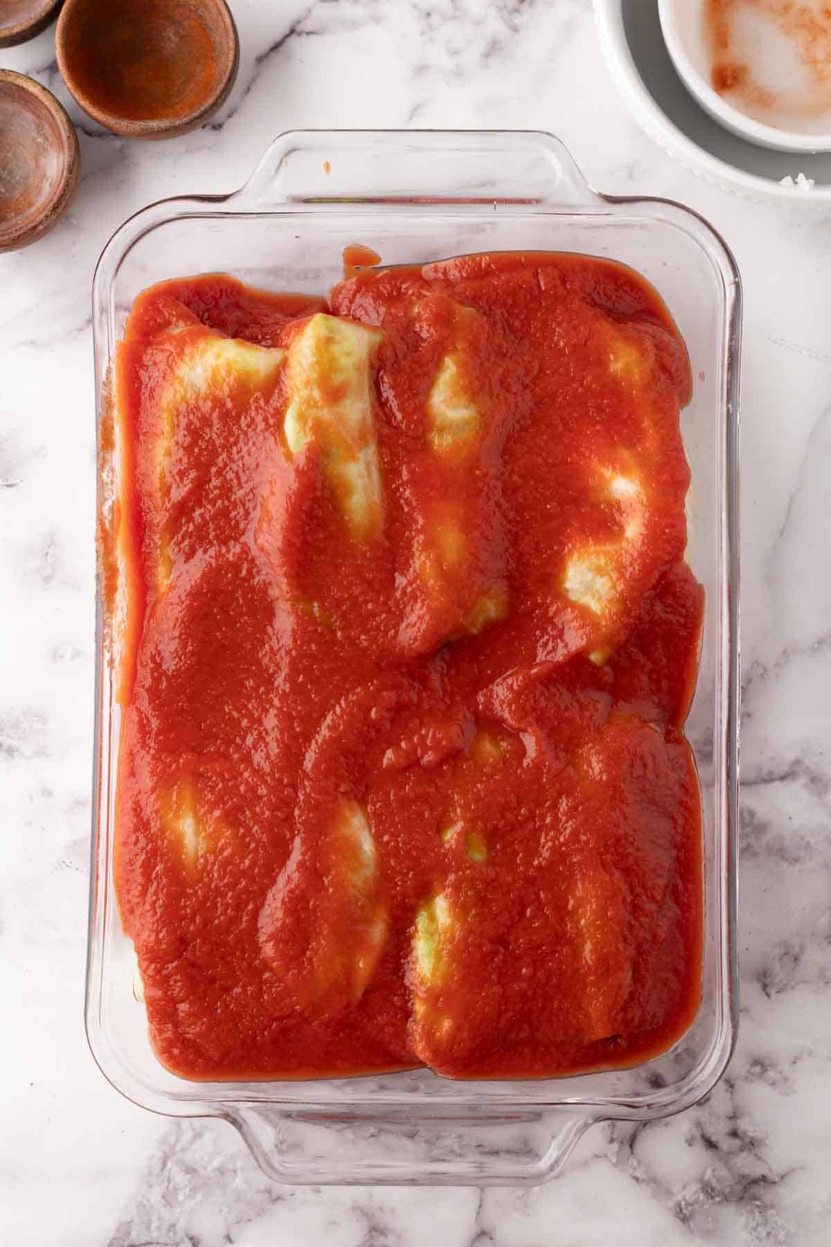 A glass baking dish filled with stuffed cabbage rolls covered in red tomato sauce, sits on a white marble surface with small empty bowls nearby.