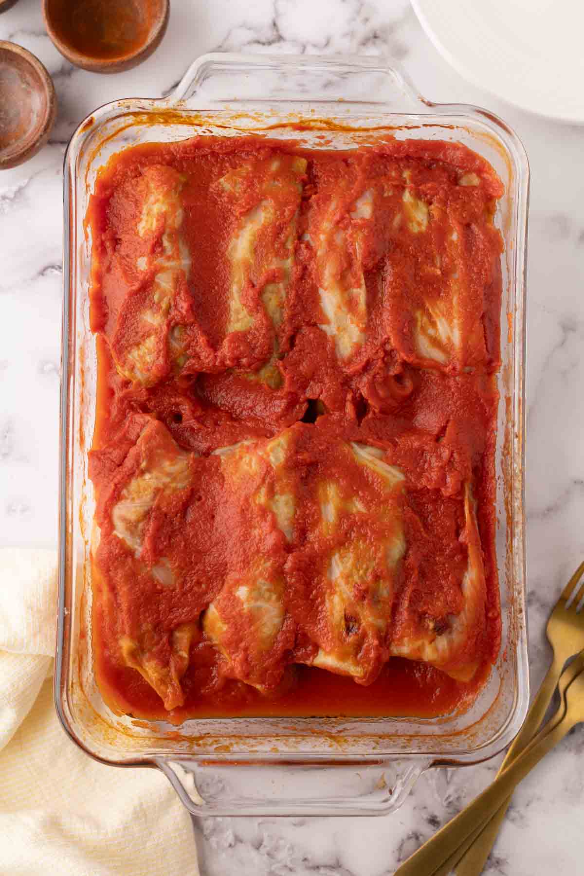 A glass baking dish filled with a stuffed cabbage rolls recipe, covered in tomato sauce, sits on a marble countertop next to a yellow napkin and gold utensils.