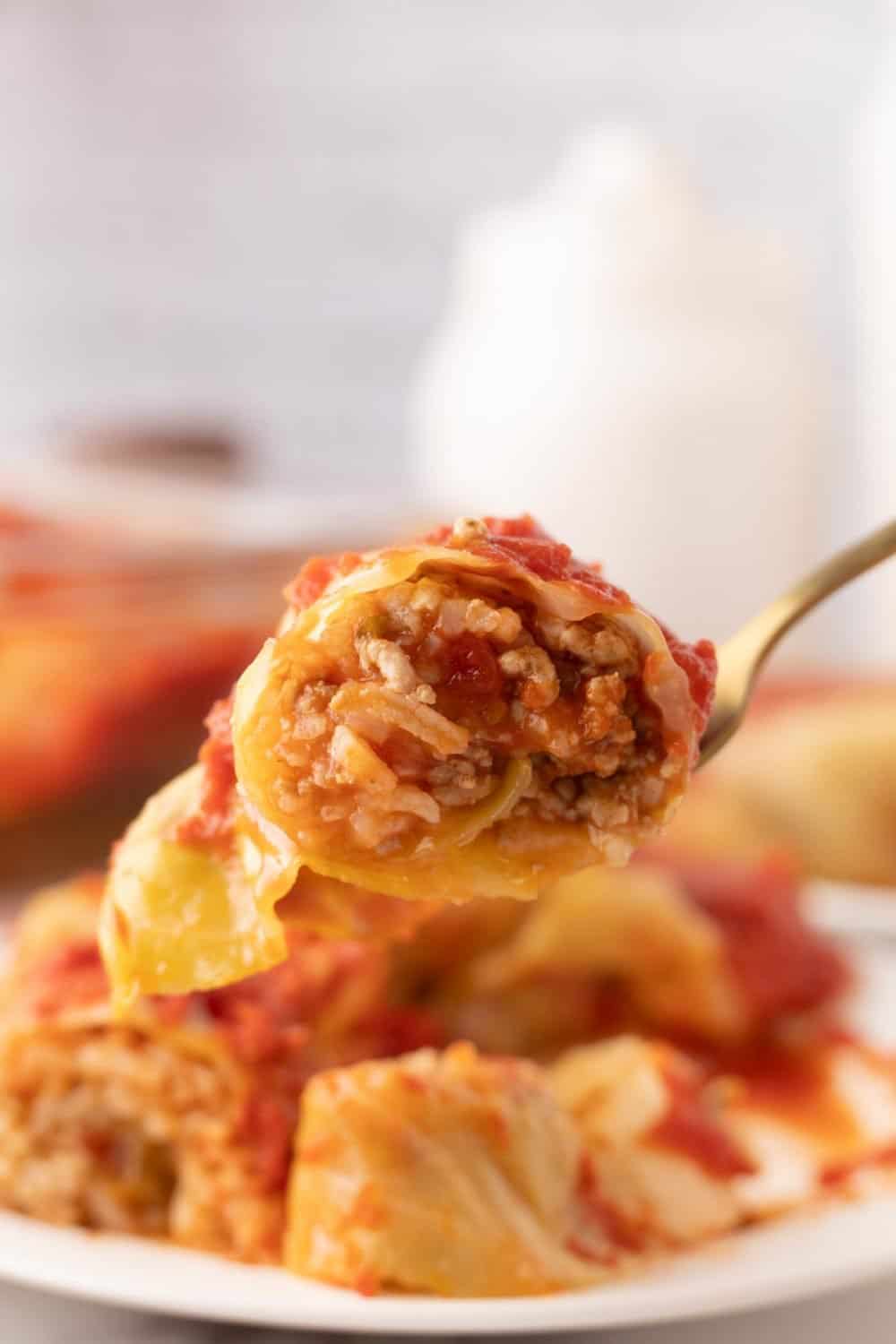 A close-up of a fork holding a bite of stuffed cabbage roll filled with rice and ground meat, topped with tomato sauce, with more cabbage rolls on a plate in the background—perfect for showcasing your favorite stuffed cabbage rolls recipe.