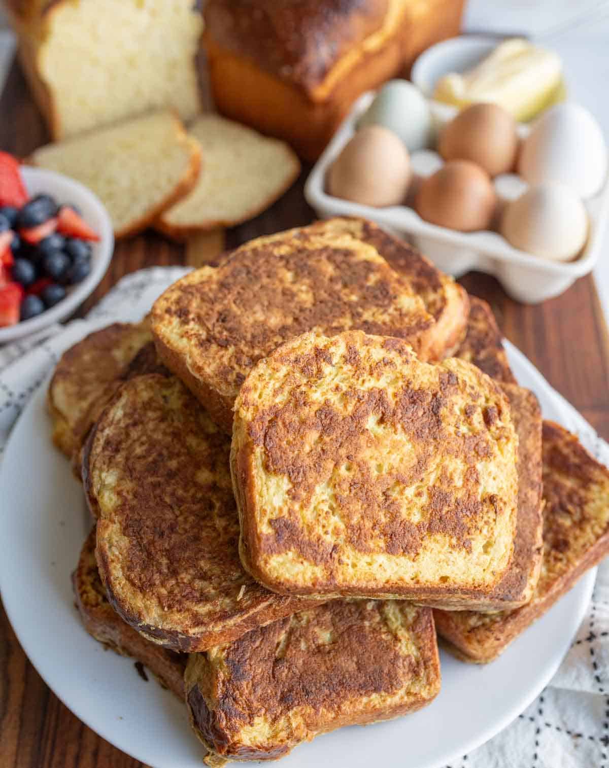 A plate of golden-brown brioche French toast slices sits on a table. In the background are eggs, butter, mixed berries in a bowl, and sliced bread, evoking a cozy breakfast scene perfect for any brioche French toast recipe.