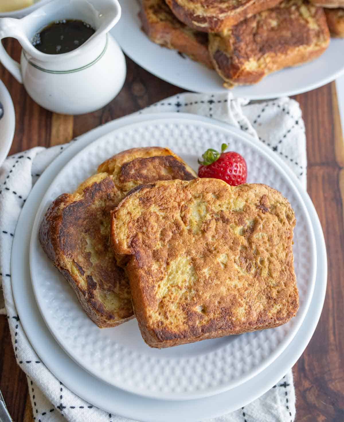 Two slices of golden-brown brioche French toast are served on a white plate, garnished with a fresh strawberry. A pitcher of syrup and extra brioche French toast recipe await in the background atop a white textured towel.