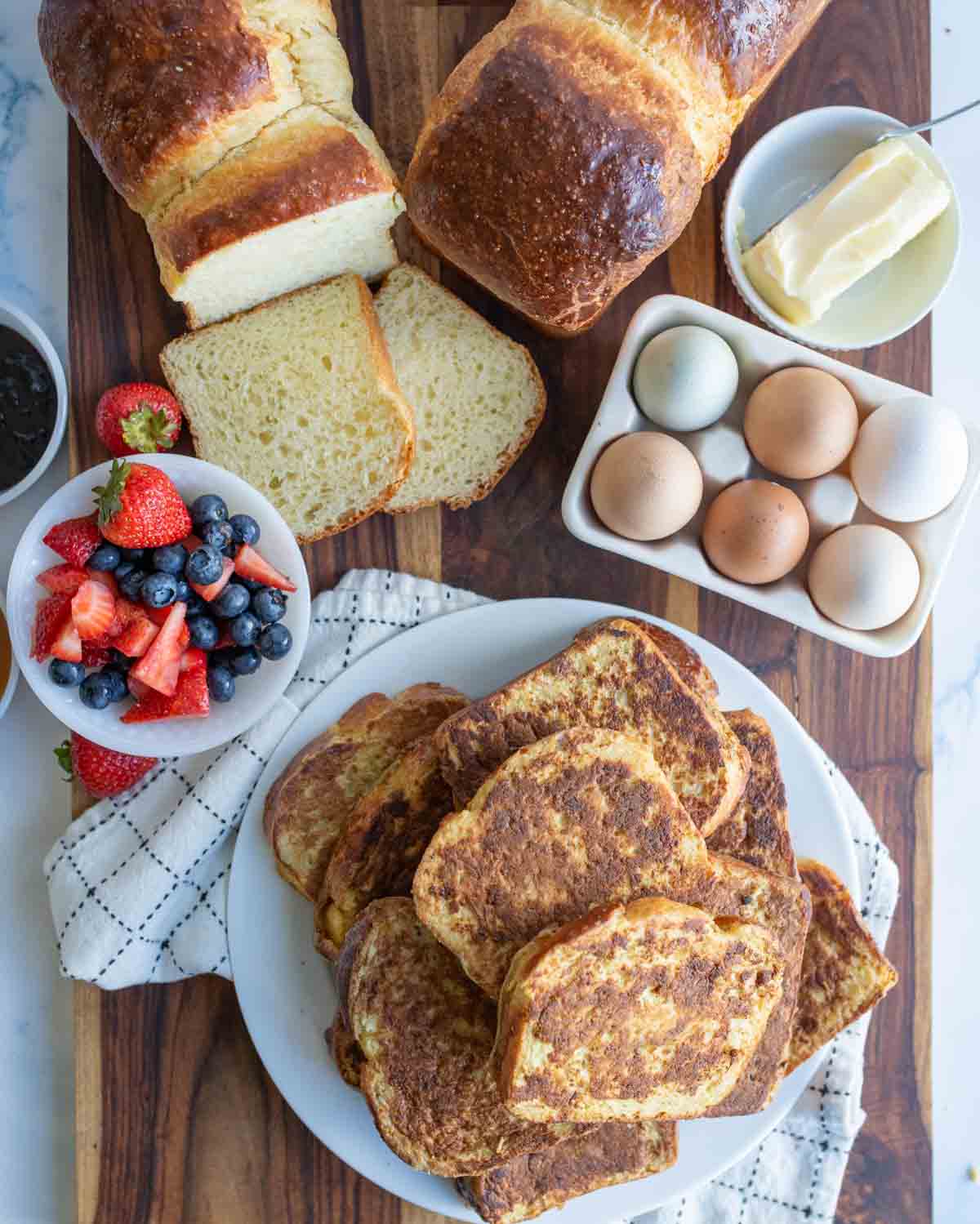 A plate of golden-brown brioche French toast sits on a wooden board, surrounded by sliced bread, a bowl of mixed berries, a dish of butter, and a carton of eggs—perfect for trying your favorite brioche French toast recipe.