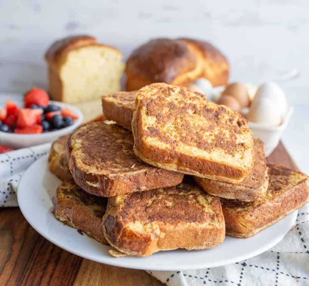A plate of golden-brown brioche French toast slices is stacked on a plate. In the background, a loaf of bread, a bowl of fresh berries, and some eggs sit on a wooden surface—perfect for this brioche French toast recipe.