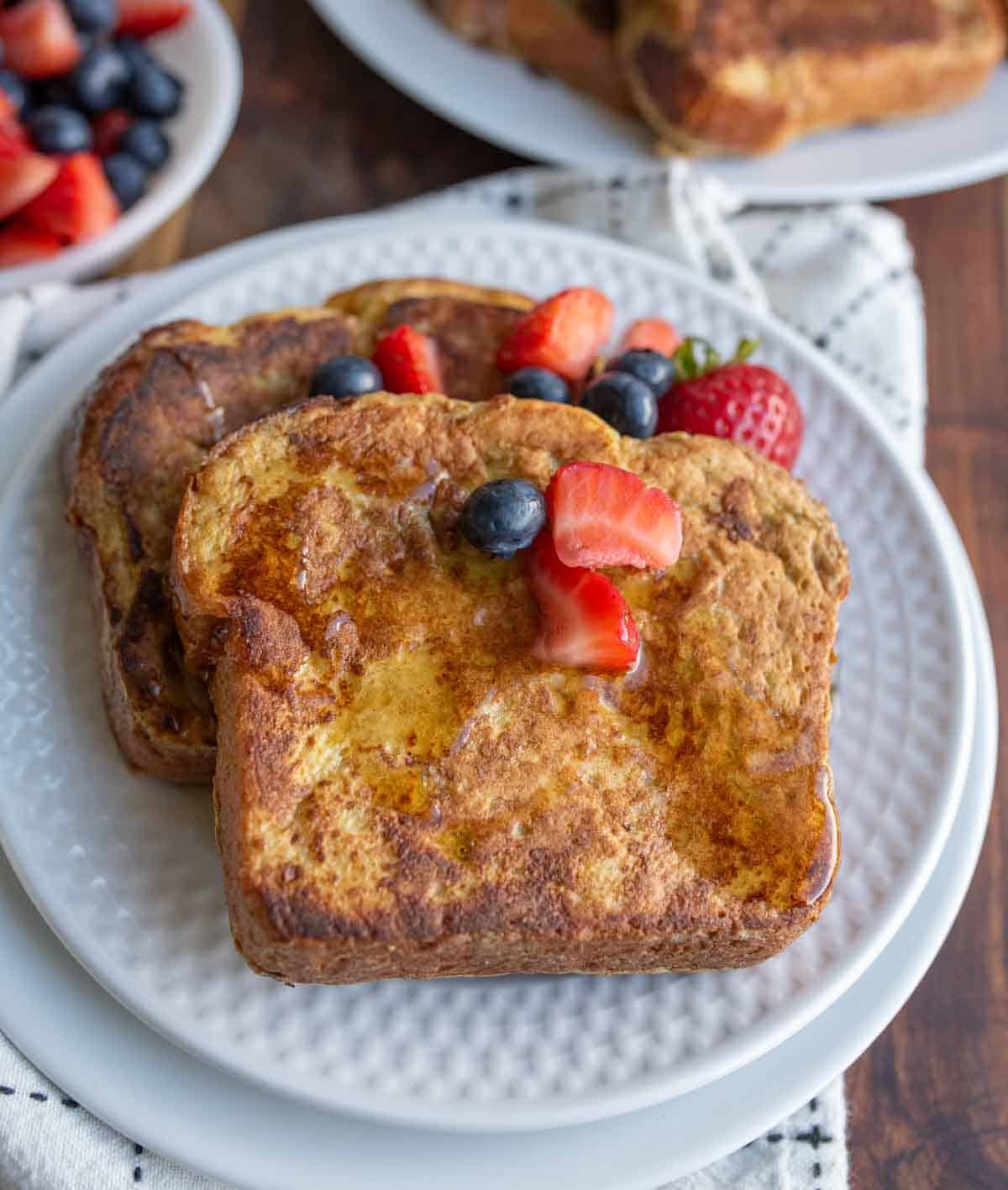 Two slices of brioche French toast topped with syrup, strawberries, and blueberries on a white plate, with a bowl of mixed berries in the background. Perfect for anyone searching for the ultimate brioche French toast recipe.