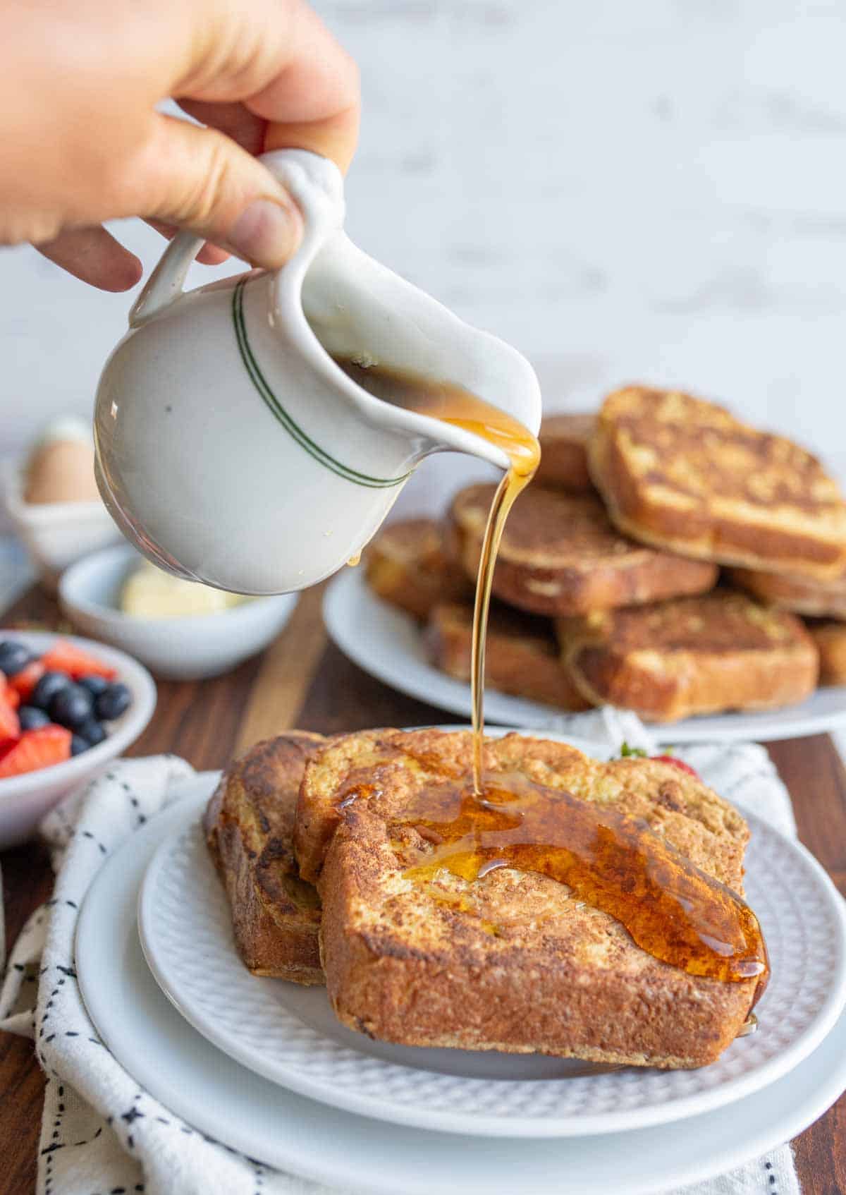 A hand pours syrup from a small pitcher onto slices of brioche French toast on a plate, with a bowl of mixed berries and a stack of more French toast from the brioche French toast recipe visible in the background.