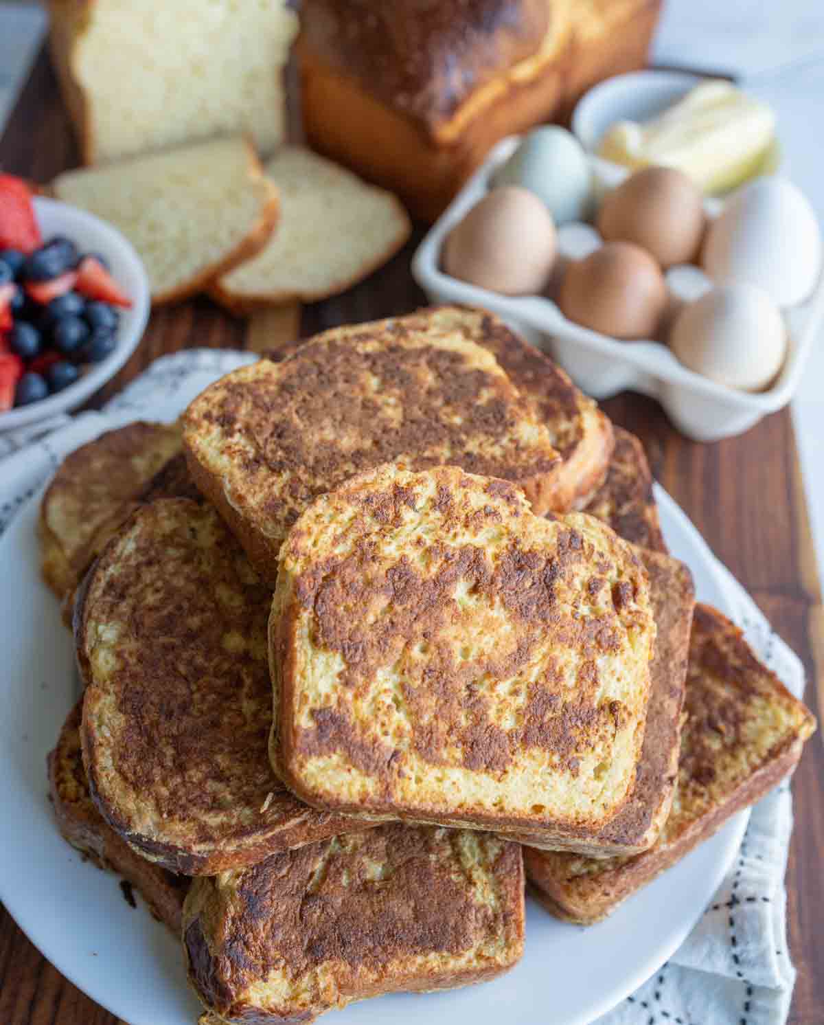 A plate stacked with slices of brioche French toast sits on a wooden board. Nearby are fresh eggs, butter, a loaf of bread, and a bowl of mixed berries—perfect for trying your own brioche French toast recipe.