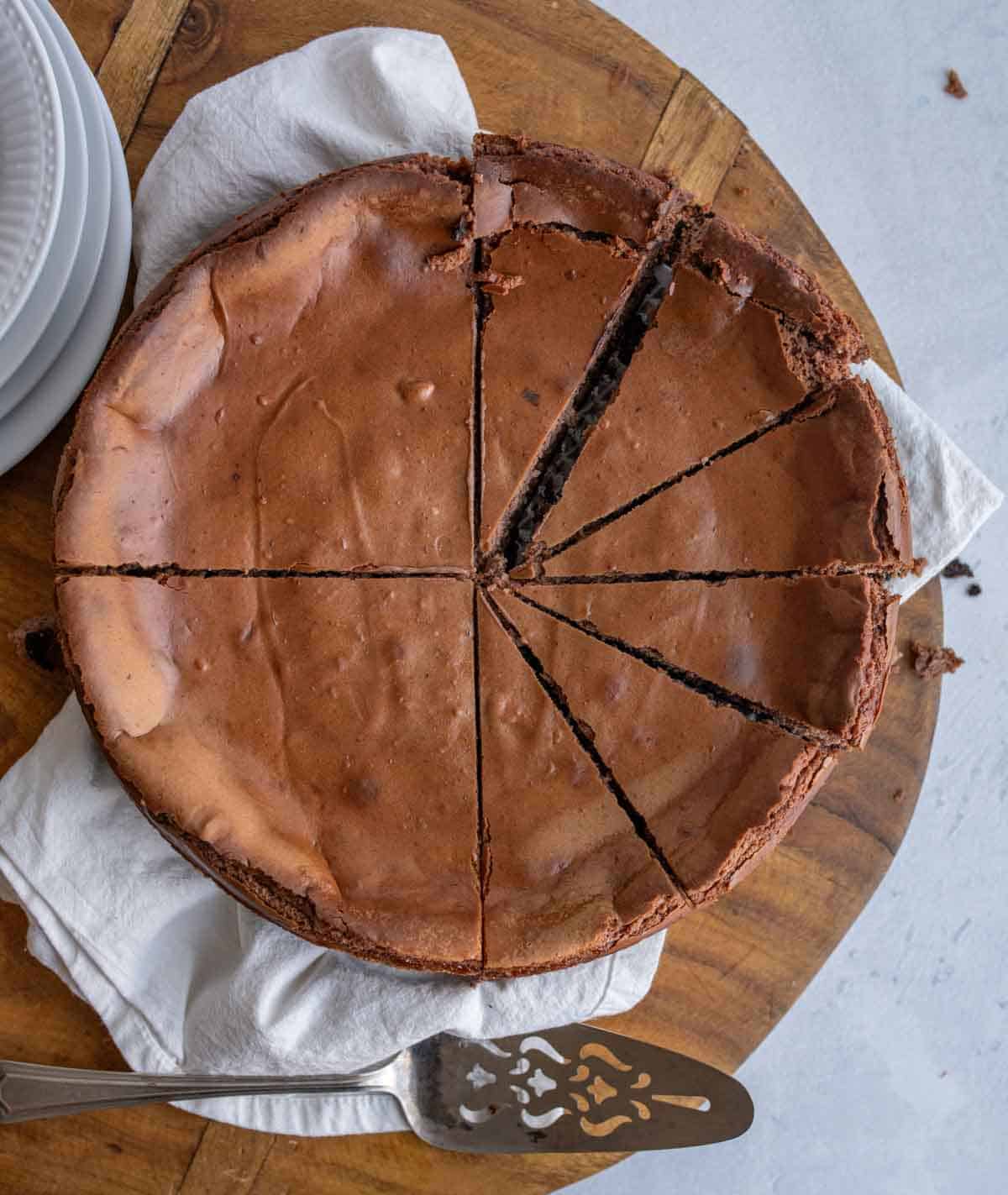 A chocolate cheesecake recipe comes to life as a decadent cake, cut into eight slices, sits on a wooden board with a white cloth underneath. A cake server and stacked white plates are placed nearby.