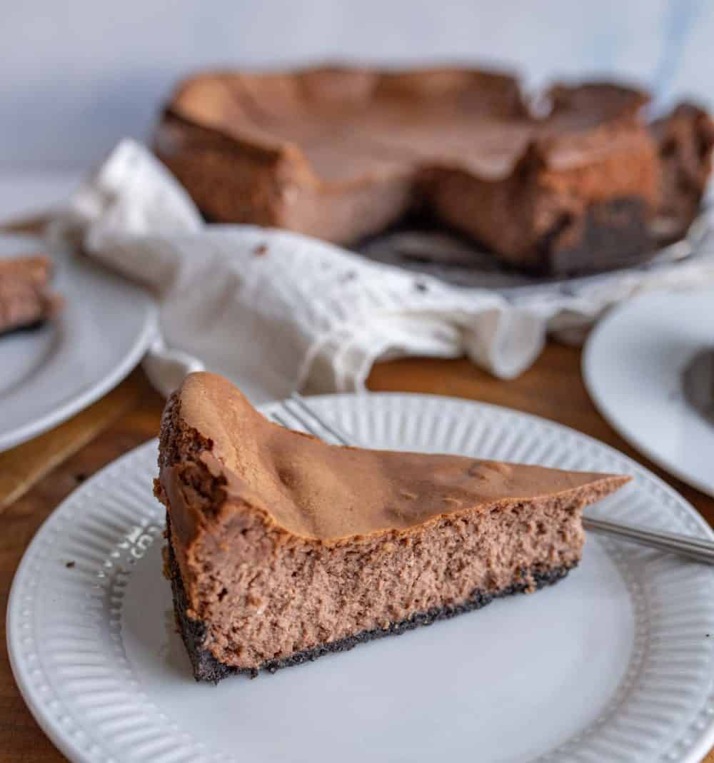 A slice of chocolate cheesecake with a dark cookie crust is served on a white plate, highlighting the rich results of this chocolate cheesecake recipe, with the remaining cheesecake and a white napkin visible in the background.