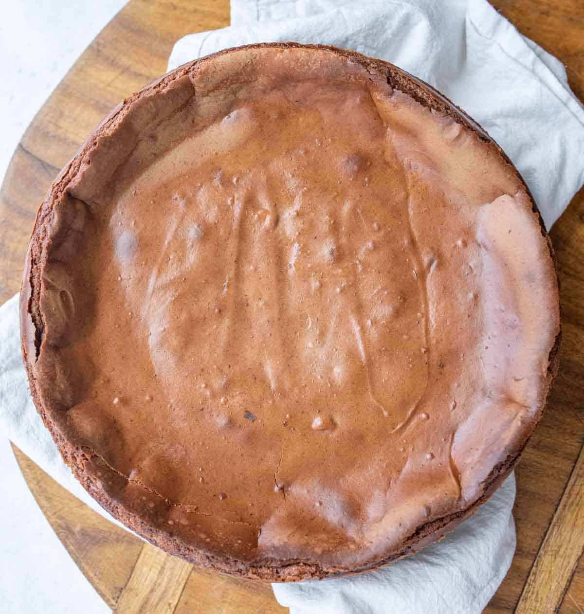 A round, rich chocolate cheesecake made from a classic chocolate cheesecake recipe sits with a smooth, slightly cracked top on a white cloth on a wooden surface.