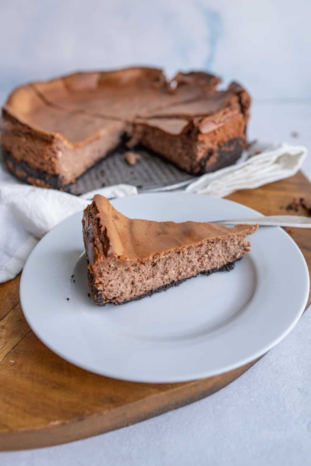 A slice of chocolate cheesecake from a classic chocolate cheesecake recipe sits on a white plate in the foreground, with the remaining cheesecake in the background on a wooden board; a fork rests nearby.
