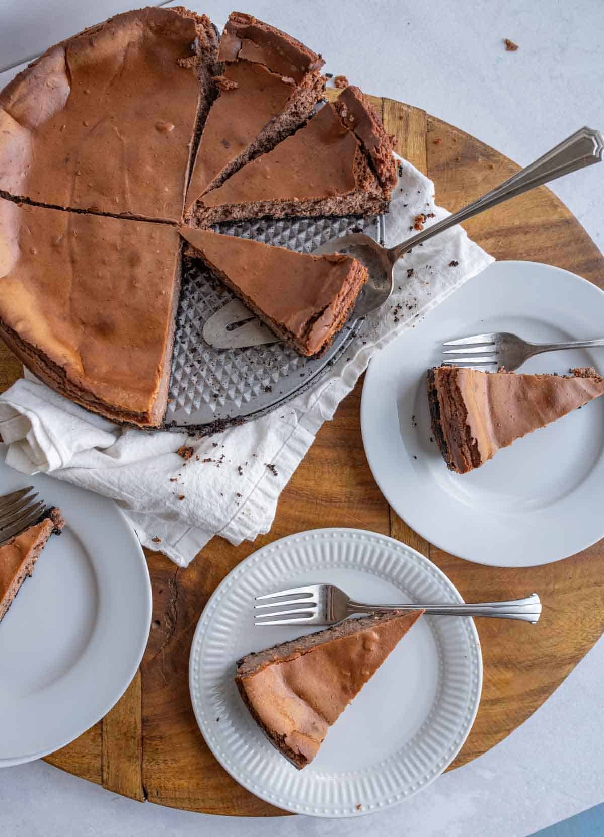 A sliced cake, made from a chocolate cheesecake recipe, sits on a round wooden board, with three slices served on white plates, each with a fork. A cake server rests beside the remaining cake.