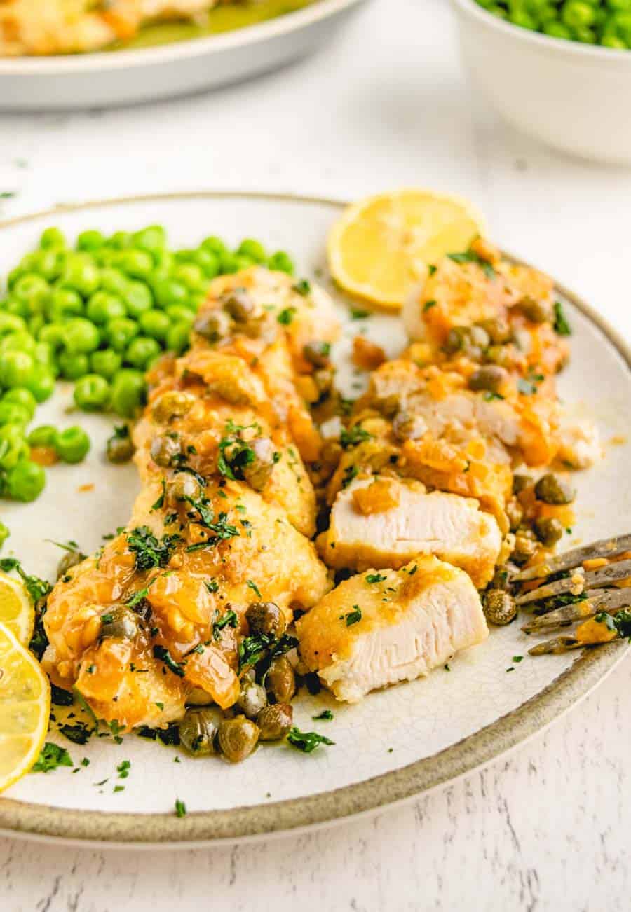 A plate with easy chicken piccata—sliced chicken breast topped with lemon-caper sauce and sprinkled with herbs, served alongside green peas and lemon slices.