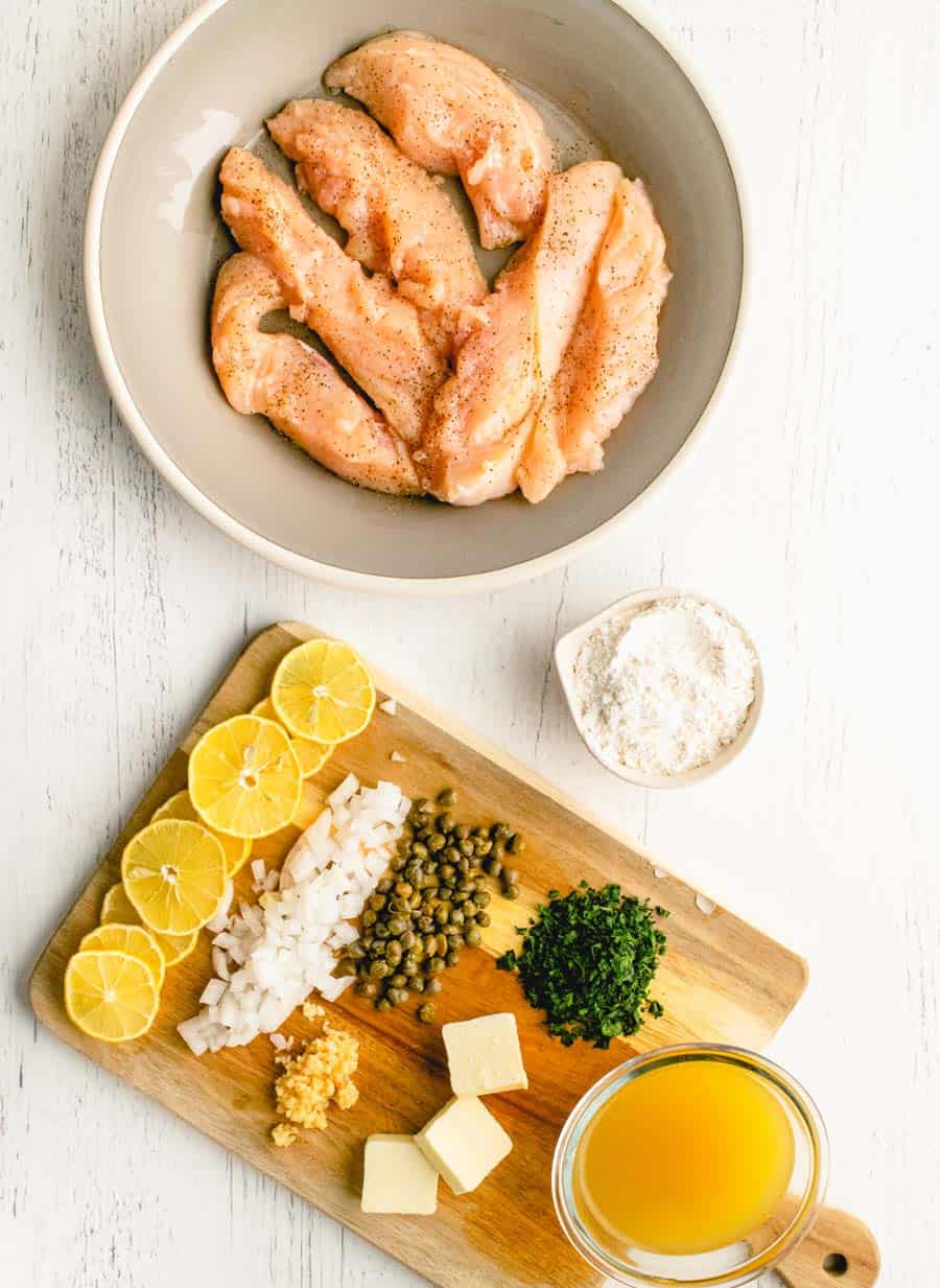 A bowl of raw, seasoned chicken tenders sits beside a cutting board with lemon slices, chopped onions, garlic, capers, parsley, butter, flour, and broth—perfect for preparing easy chicken piccata on a white surface.