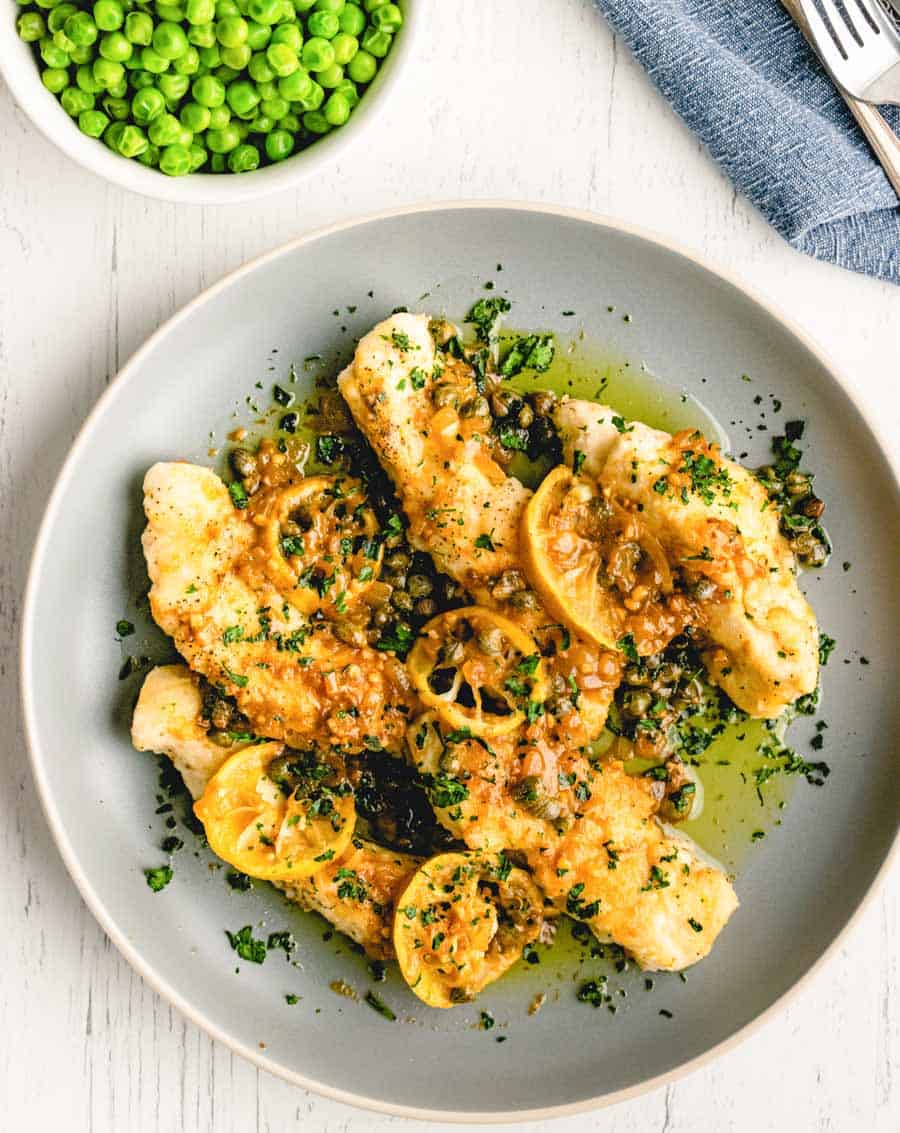 A plate of fish fillets topped with lemon slices, capers, and a light sauce—much like an easy chicken piccata—garnished with chopped herbs. A bowl of green peas sits nearby on the white table.