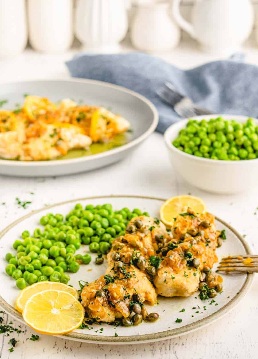 A plate with easy chicken piccata garnished with parsley and lemon slices, served alongside green peas. Another plate of food and a bowl of peas are in the background, on a white table with a blue napkin.