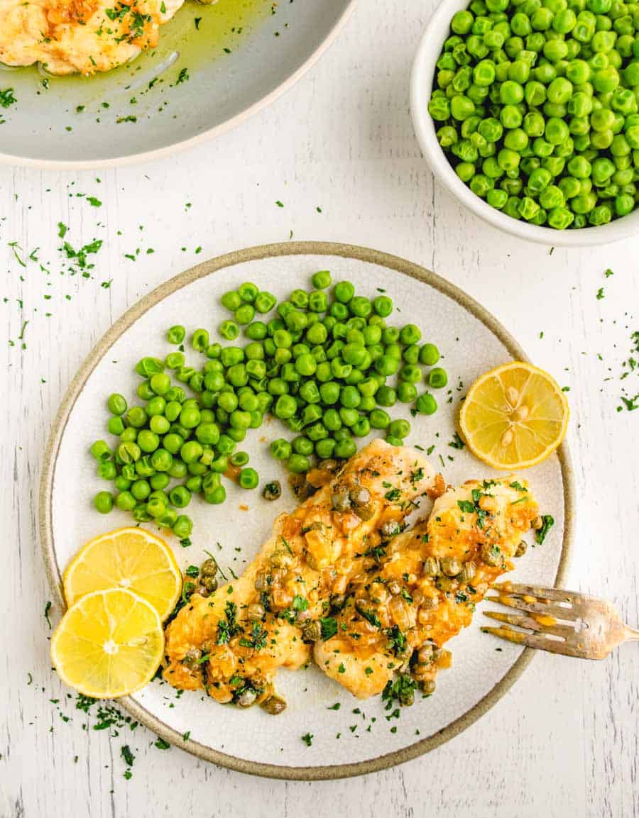 A plate with easy chicken piccata topped with capers and sauce, garnished with parsley and lemon slices, served with a side of green peas. A bowl of peas and a partial plate are visible on a white wooden table.