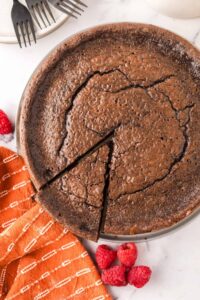 A flourless chocolate cake with a cracked top sits on a plate, one slice slightly separated. An orange patterned cloth and fresh raspberries are nearby, with a stack of plates and forks in the background—perfect for sharing this chocolate cake recipe.