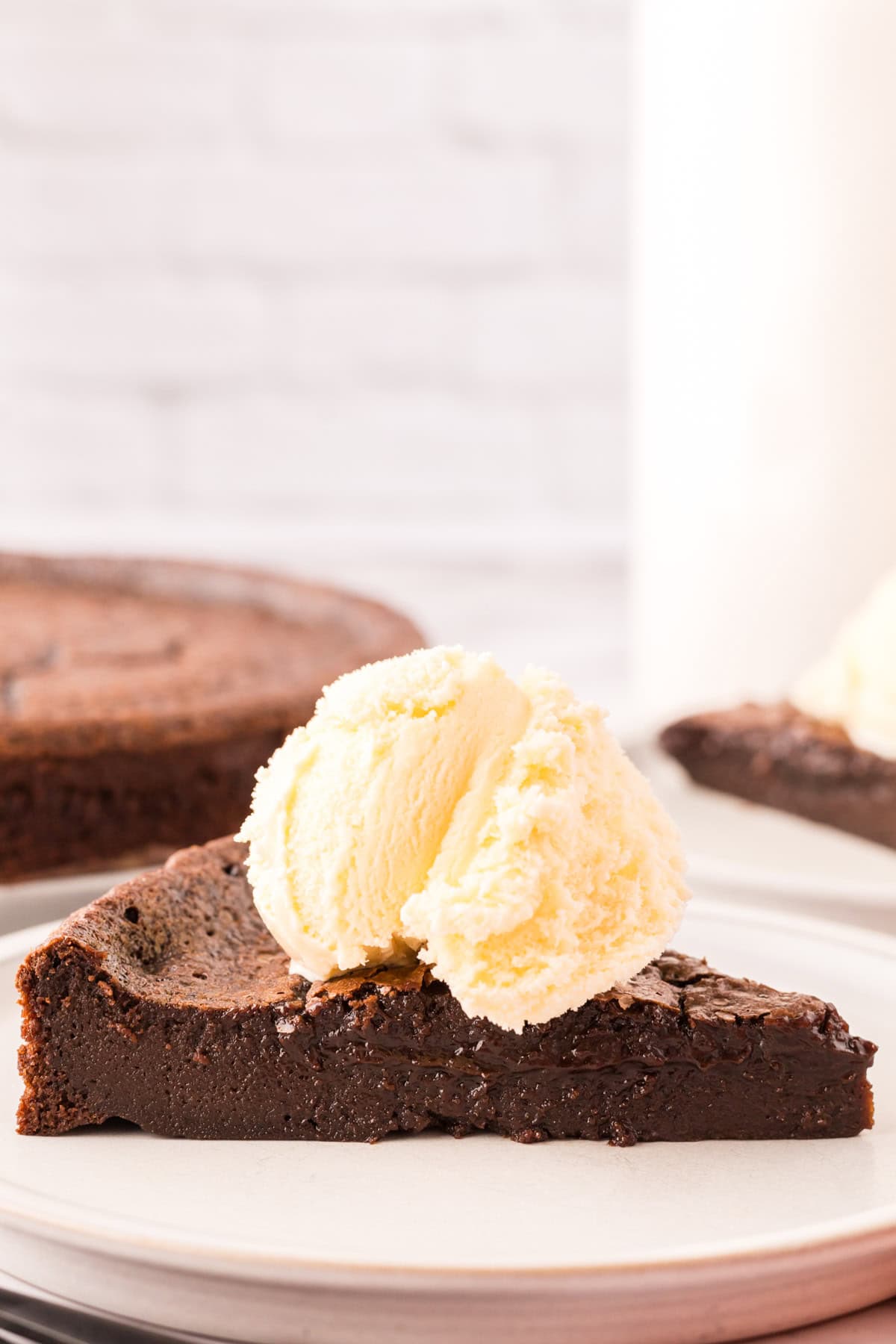 A slice of rich, fudgy flourless chocolate cake recipe topped with a scoop of vanilla ice cream, served on a white plate with another slice and cake in the background.