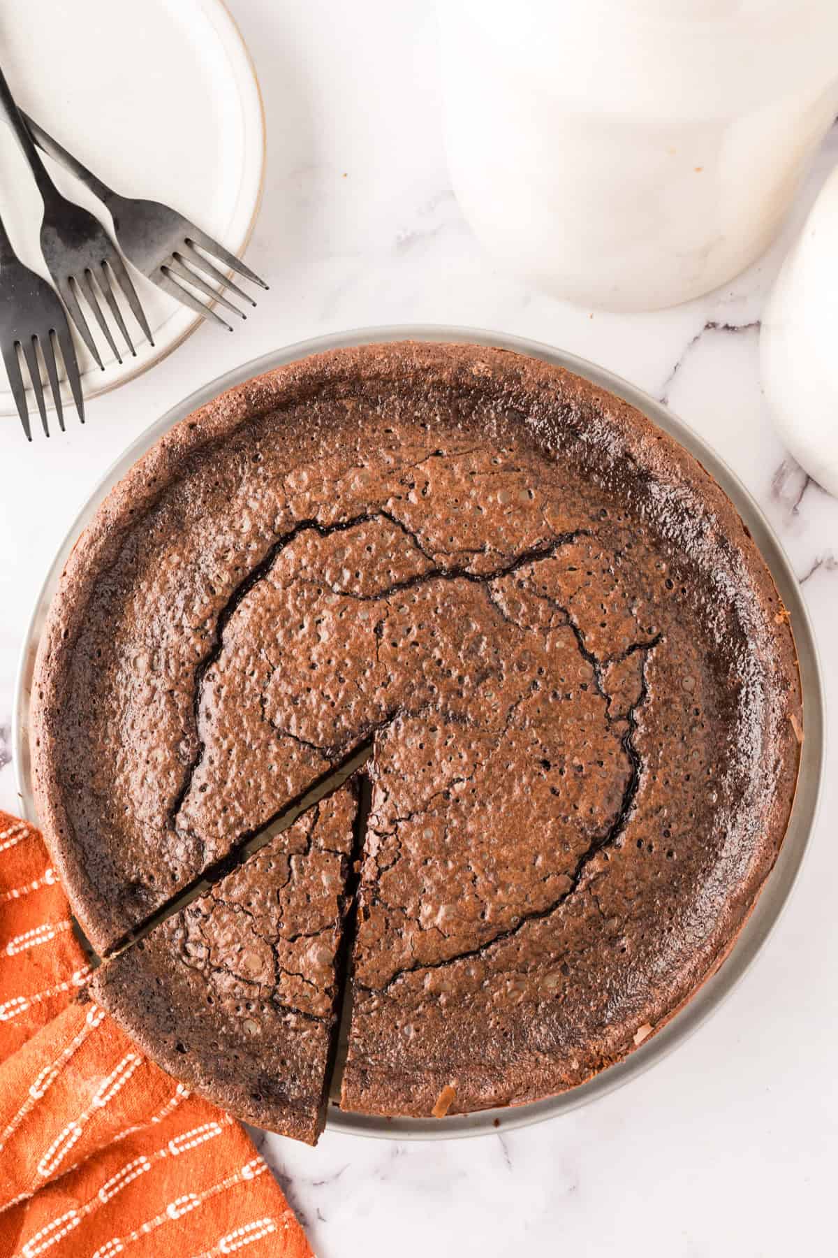 A whole, crackly-topped flourless chocolate cake recipe sits on a marble surface next to an orange cloth, three black forks, and a white plate—one slice partially removed.