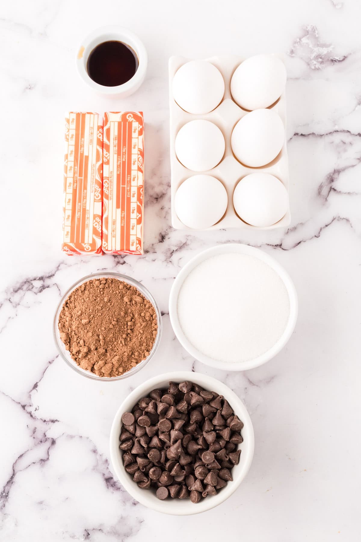 Baking ingredients on a marble surface for a flourless chocolate cake recipe: sticks of butter, a small bowl of vanilla extract, six eggs in a carton, a bowl of cocoa powder, granulated sugar, and chocolate chips.