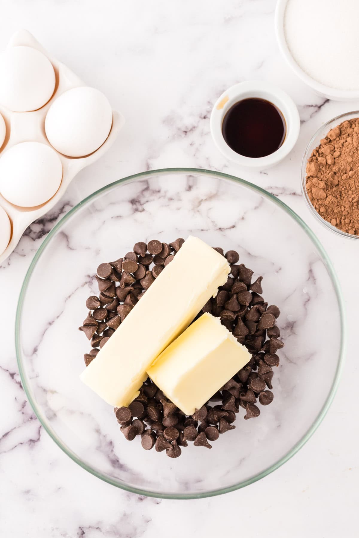 A glass bowl with chocolate chips and two sticks of butter sits on a marble surface—essential ingredients for a flourless chocolate cake recipe. Nearby are eggs in a carton, vanilla extract, sugar, and cocoa powder ready to be mixed.