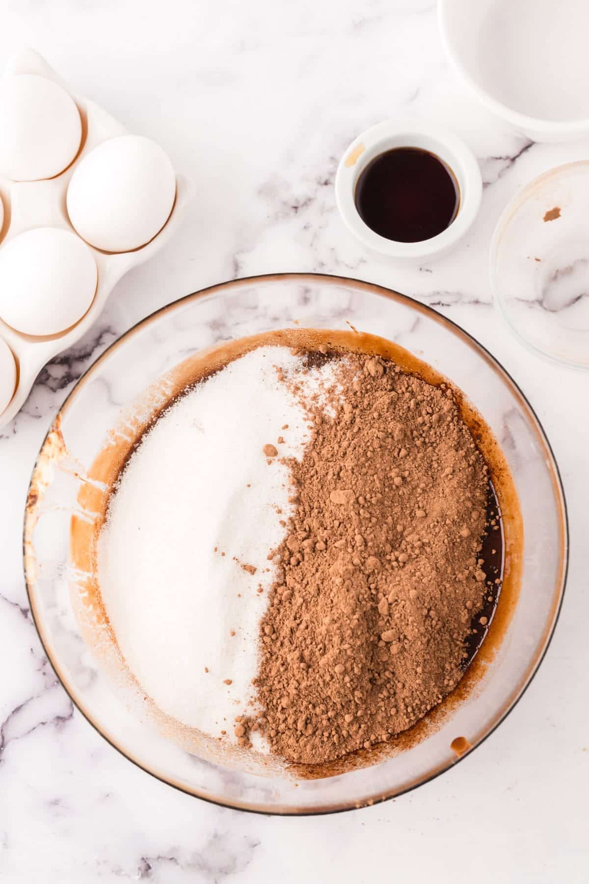 A glass bowl filled with white sugar and cocoa powder sits on a marble counter—key ingredients for a flourless chocolate cake recipe. Nearby are eggs in a carton, a small bowl of vanilla extract, and an empty white bowl.